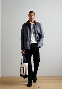 Gray waterproof jacket with brown collar, white shirt, and black pants. Holding a beige and navy tote bag. Black shoes. Wood floor background.