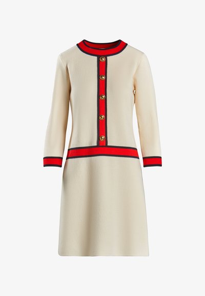 Bézs színű, térd hosszúságú ruha, hosszú ujjakkal, piros és tengerészkék kiegészítőkkel, kerek nyakkal és arany gombokkal az elején. Simább textúra.