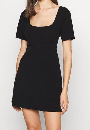 Woman wearing a short black dress with a square neckline and short sleeves against a plain white background.