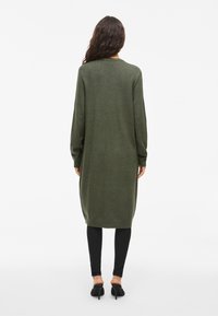 Long gilet en tricot olive avec une coupe décontractée, des poignets côtelés et une fente sur le côté, associé à un legging noir et des chaussures à bout ouvert.