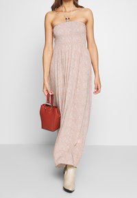 Axellös maxiklänning med blommönster i ljusrosa, tillverkad av mjukt tyg. Kombinationen med en liten röd handväska och beige klackstövlar.