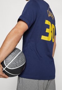 Camiseta de algodón azul marino con el texto amarillo "CURRY" y el número "30" impresos en la parte posterior, combinada con pantalones cortos grises y sosteniendo un baloncesto negro texturizado.