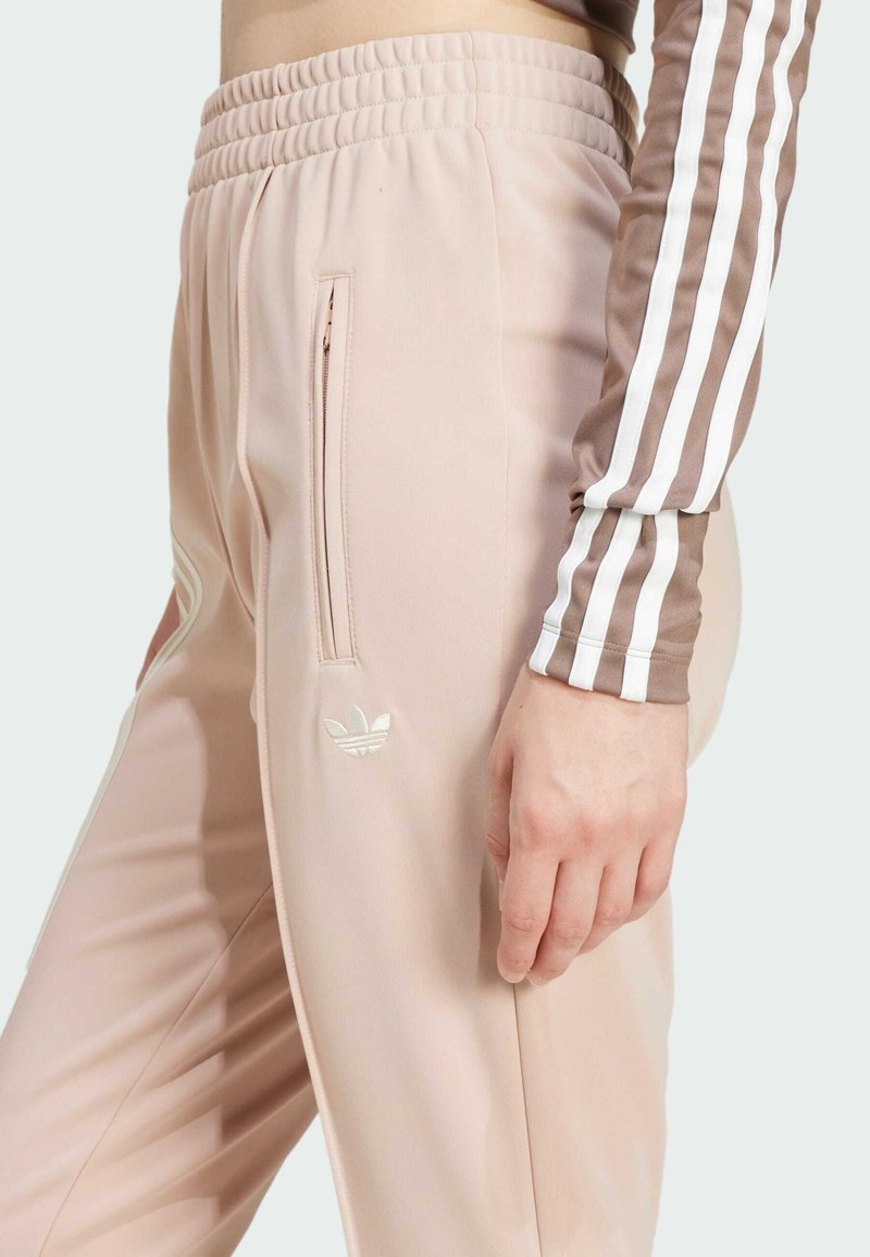 Jogging en rose clair avec une taille côtelée, une poche zippée sur le côté et un logo brodé. Présente des rayures blanches sur le côté pour l'accent.