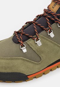 Olive green buty trekkingowe z teksturowanej zamszowej i nylonowej siateczki, czapka z czarnej gumy na palcach, pomarańczowe akcenty na podeszwie oraz metalowe oczka na sznurówki.