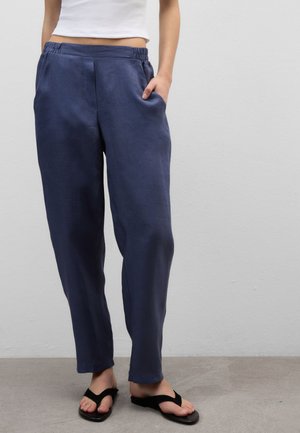 Person trägt marineblaue, locker sitzende Hose, weißes kurzes Top und schwarze Flip-Flops, steht mit einer Hand in der Tasche.