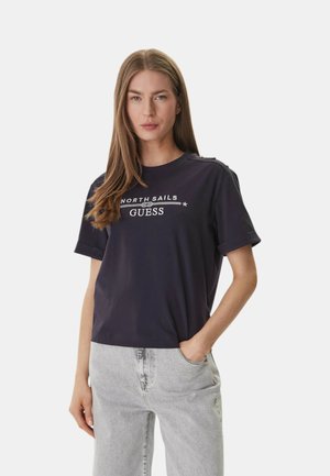 Vrouw met lang haar, gekleed in een zwart "North Sails GUESS" T-shirt en lichtgrijze jeans, met één hand in de zak, staand tegen een witte achtergrond.
