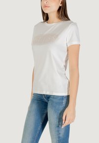 Camiseta blanca de algodón con mangas cortas y un escote redondeado, adornada con un logo "GUESS" brillante en un diseño texturizado.