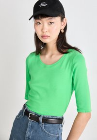 T-shirt à manches longues, ajusté, en coton vert vif avec un col rond, associé à un jean en denim taille haute et une ceinture noire.