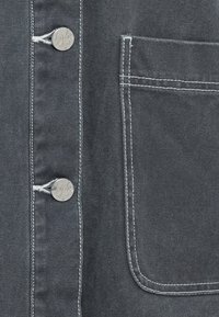 Tissu gris foncé avec des détails de couture visibles, boutons en métal, et une poche sur le côté droit, mettant en avant un design textile robuste.