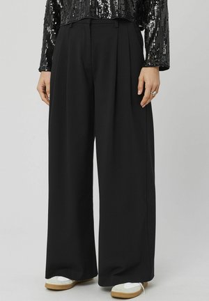 Pantalon noir à jambes larges en tissu doux, avec plis sur le devant, taille haute et texture lisse. Porté avec des baskets blanches.