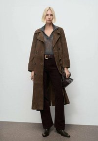 Abrigo trench de ante marrón, suéter gris de cuello en V, pantalones de pana marrón oscuro y botas de tobillo negras. Lleva una bolsa marrón oscura.