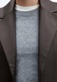 Detailaufnahme einer Person, die eine braune Lederjacke über einem grauen Pullover mit einem weißen Rundhalsshirt darunter trägt.