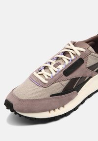 Baskets en daim beige et mauve avec tissu, lacets crème, semelle noire et bandes latérales noires, inclinées pour montrer les détails avant et latéraux.