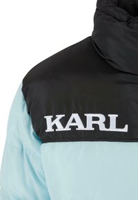 Polstret jakke med en sort øverste sektion og lyseblå nederste del. Fremtrædende hvid "KARL" logo broderet på brystet.
