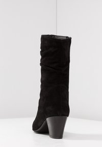 Botte en daim noire mi-mollet au design flou et talon moyen empilé, présentée sur une surface blanche contre un fond uni.