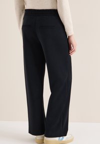 Pantalon large noir à taille élastique, doté de deux poches arrière, porté avec un pull clair et des baskets blanches.