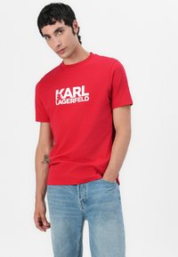 Raudona medvilninė marškinė su baltu "KARL LAGERFELD" logotipu; trumpomis rankovėmis ir apvaliu kaklu; derinami su šviesiai mėlynomis džinsomis.