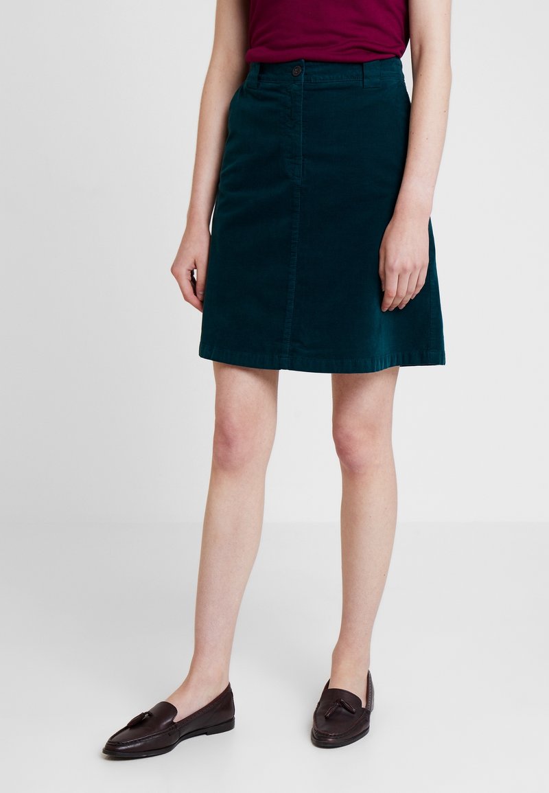 Jupe A-line en velours côtelé teal avec une fermeture à boutons à l'avant et des poches latérales, tombant au-dessus du genou, portée avec des mocassins foncés.