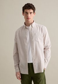 Camisa de botón de color beige claro con rayas verticales blancas, un bolsillo en el pecho y cuello, combinada con pantalones de color verde oliva.