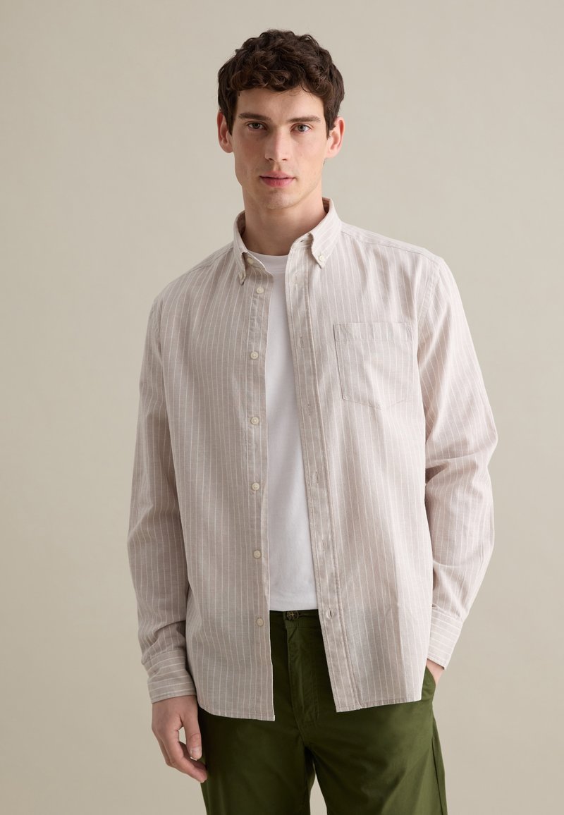 Camisa de botón de color beige claro con rayas verticales blancas, un bolsillo en el pecho y cuello, combinada con pantalones de color verde oliva.