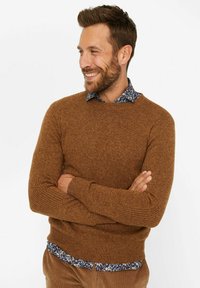 Brauner strukturierter Pullover mit Rundhalsausschnitt, kombiniert mit einem blauen, blumengemusterten Hemd, das darunter hervorschaut.