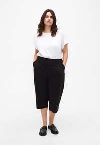 Pantalons noirs coupés avec poches latérales, associés à un t-shirt blanc ample. Le mannequin porte des chaussures noires à enfiler à texture.