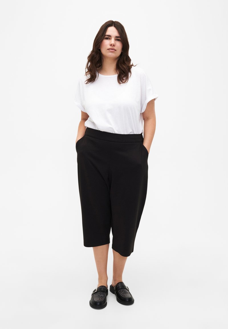 Pantalons noirs coupés avec poches latérales, associés à un t-shirt blanc ample. Le mannequin porte des chaussures noires à enfiler à texture.