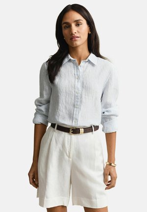 Femme portant une chemise à rayures bleu clair à boutons avec les manches retroussées, un short blanc taille haute, une ceinture foncée et des bijoux dorés.