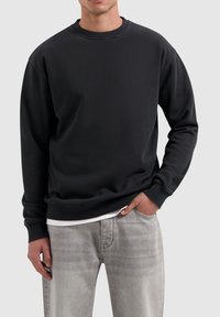 Zwarte crewneck sweatshirt met ribgebreide manchetten en zoom. Zachte stof, casual pasvorm en zichtbare witte onderlaag bij de zoom, gedragen met lichtgrijze jeans.