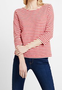 Rot-weiß gestreiftes Langarmshirt mit einem Bootsausschnitt. Weicher Stoff, gerippte Struktur, leicht lockere Passform, kombiniert mit dunklen Jeans.