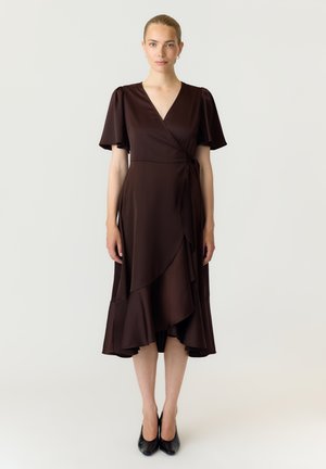 Femme aux cheveux blonds portant une robe portefeuille marron foncé avec des manches volantées et des talons pointus noirs, debout devant un fond clair uni.