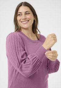Leende kvinna som justerar manschetten på en långärmad lavendelfärgad tröja med ribbade och veckade texturerade ärmar framför en neutral bakgrund.