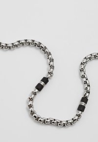 Chaîne en métal argenté avec des maillons ronds, rehaussée de caoutchouc noir et de détails métalliques, formant un design en boucle continue.