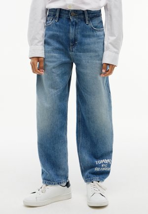 Persona che indossa jeans blu larghi con il testo "Tommy NYC Hilfiger" vicino alla caviglia sinistra, sneaker bianche e una maglietta bianca a maniche lunghe.