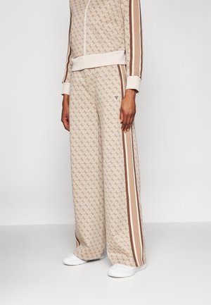 Beige bukser med vide ben og geometrisk mønster med brune striber, båret med matchende jakke og hvide sneakers på en stående person.