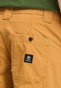 Tan arbejdsshorts med en baglomme, der har et sort Timberland logo-patch og en knaplukning. Tyk bomuldsstof med en glat tekstur.