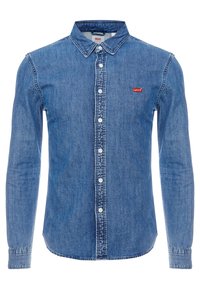 Sininen farkkupaita pitkillä hihoilla, nappikaulus, valkoiset napit ja pieni punainen Levi's-logo rinnassa. Pehmeä materiaali, klassinen istuvuus.