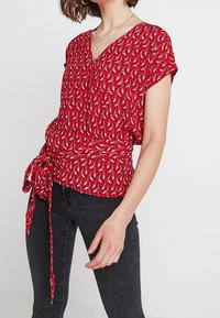 Červený top s krátkými rukávy a výstřihem do V, s květinovým vzorem, uvázaným pasem a volným střihem. Vyrobeno z lehké, texturované tkaniny.