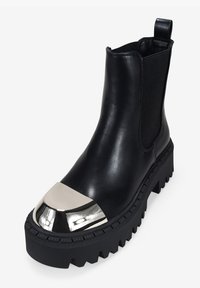 Botín de cuero negro hasta el tobillo con una suela gruesa negra, puntera metálica plateada, paneles laterales elásticos y una tira en la parte trasera.