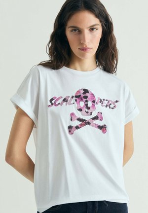 Mujer con una camiseta blanca de mangas remangadas que presenta un gráfico de calavera y huesos cruzados en estampado de leopardo rosa con la palabra "SKALOPPERS".