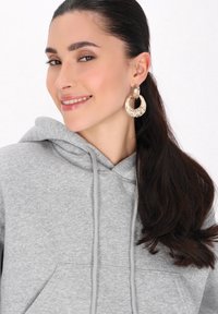 Felpa grigia con cappuccio, dotata di una tasca frontale. Primo piano di un modello che indossa orecchini a cerchio in oro con texture e capelli lunghi e scuri.