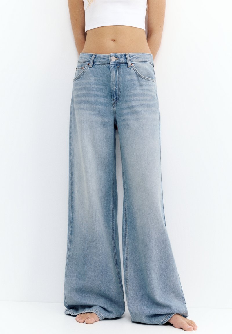 Personne portant un jean large bleu clair et un crop top blanc, debout pieds nus contre un mur blanc.