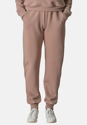 Person trägt hellbraune Jogginghosen und weiße Sneakers, Hände in den Taschen, steht vor einem einfarbigen Hintergrund.