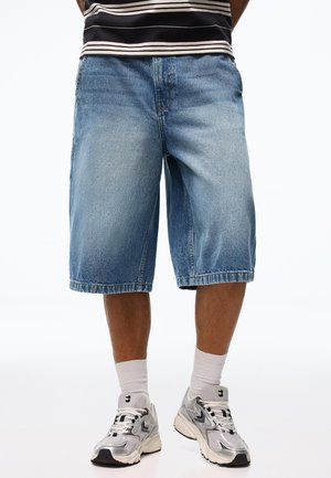 Férfi kék farmer térdig érő rövidnadrágban, fehér zokniban és ezüst sportcipőben, egyszínű háttér előtt áll.