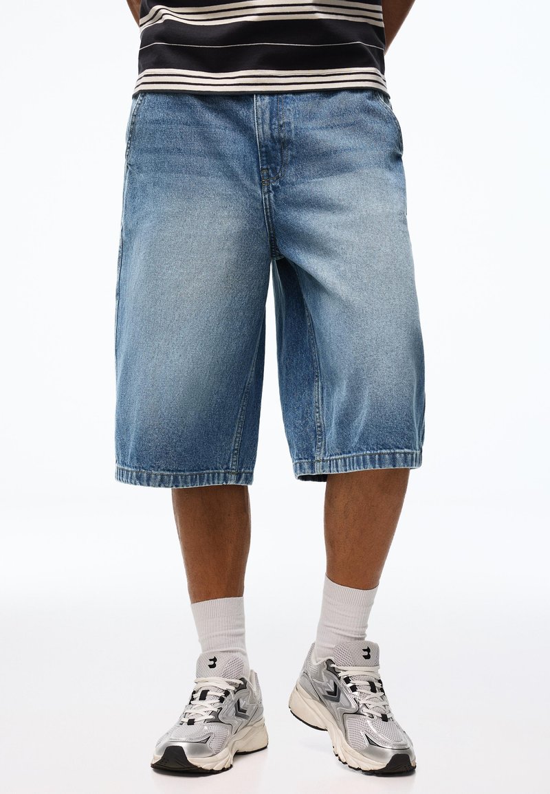 Mann trägt blaue Denim-Knielange Shorts, weiße Sportsocken und silberne Turnschuhe, steht vor einem schlichten Hintergrund.
