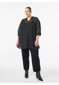 Kvinde iført en sort gennemsigtig bluse med prikker, sorte bukser og sorte spidse ankelstøvler med sølvnitter, stående mod en hvid baggrund.