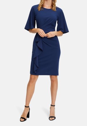 Femme portant une robe bleu marine longueur genou avec des manches volantées et un détail de volants en cascade à l'avant, associée à des escarpins noirs à bride à la cheville.