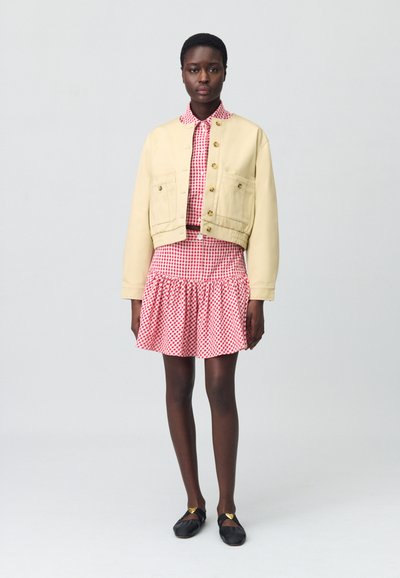 Veste beige courte avec des boutons dorés, associée à une robe à carreaux rouges avec un ourlet volanté. Ballets noirs avec des accents dorés.