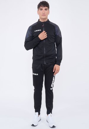 Mann trägt schwarzen LEGEA Trainingsanzug mit geometrischem Muster auf den Schultern und weißen Sneakers, steht vor einem schlichten weißen Hintergrund.