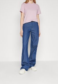 Różowa koszulka z bawełny z okrągłym dekoltem, zestawiona z niebieskimi dżinsami rozszerzającymi się u dołu oraz białymi trampkami. Materiały są gładkie i casualowe.